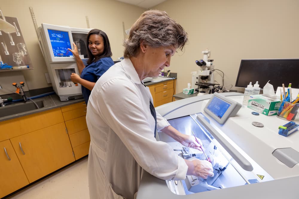 Two laboratory staff operating histology equipment in a medical lab, with microscopes, workstations, and specimen-processing instruments used for tissue analysis.